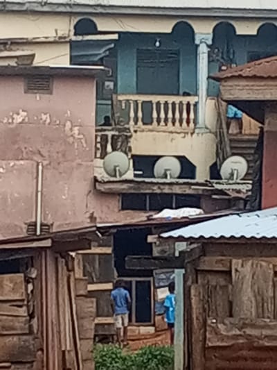 A crowded, urban scene shows dense housing with a mix of architectural styles. Buildings vary in color and construction, with some displaying weathered exteriors. Three satellite dishes are visible on one structure, along with a small balcony. Two people stand in the foreground amidst the clustered houses, adding a sense of scale to the urban environment.