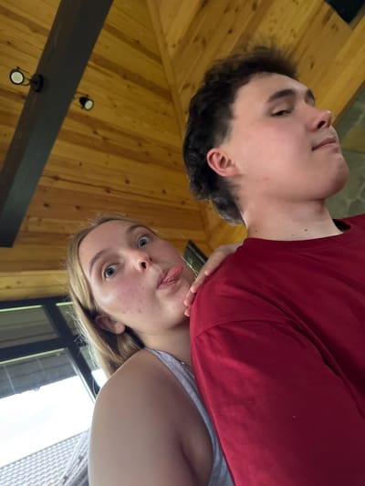In the image, a young woman with blonde hair is sticking her tongue out at the camera, her eyes wide and playful. She is leaning in from the left side of the frame, her arm resting on the shoulder of a young man who is facing away from the camera. The man is wearing a red t-shirt and has dark, curly hair. They are positioned in an indoor setting with a wooden ceiling and exposed beams, which suggests a rustic or cabin-like environment. The lighting appears to be natural, possibly from a window or skylight, casting a soft glow on their faces. The overall impression is of a casual and fun moment captured between two people. The location is identified as Ryabinino, Russia.