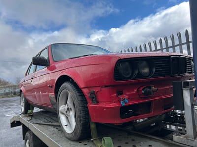 A red car is upside down and strapped to a trailer. The trailer is parked outdoors in what appears to be a parking lot or industrial area. The sky is visible in the background, with white clouds against a blue sky. A fence is partially visible between the car and the sky. The car's front end is visible, including headlights and the front bumper. One wheel is visible, partially obscuring the rear of the car. No people are visible in the image. The image was taken in the city of Shotts, United Kingdom.