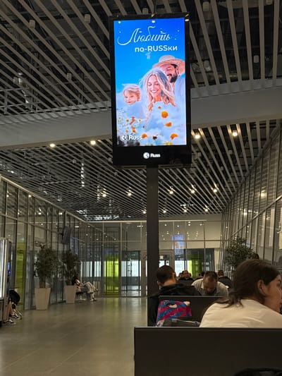 The image captures an indoor scene, likely a modern airport or public transit station, featuring a large digital advertisement screen mounted on a dark gray pillar. The screen displays a vibrant advertisement with Russian text at the top that translates to "Love." Below this text is "po-RUSSki," which can be interpreted as "in Russian style" or "like Russians." The advertisement features a smiling couple and a child surrounded by white and orange flowers. The brand logo "Russ" appears twice on the screen.

The environment is characterized by a high ceiling with a grid of diagonal white beams and numerous circular light fixtures. The floor is tiled in a light gray. Large glass windows line the right side of the image, revealing a glimpse of an exterior or another section of the building, with green and yellow lighting visible in the distance.

Several people are seated in a waiting area in the foreground and midground. In the foreground, a woman with dark hair is looking to the right, wearing a white t-shirt. To her left, a man with short hair is seated, with a backpack featuring a colorful, abstract pattern placed behind him. Further in the midground, other individuals are seated, some appearing to be sleeping or resting. Two large potted plants with dark green foliage stand near the glass windows, adding a touch of greenery to the otherwise modern interior. The overall atmosphere suggests a moment of waiting or transit within a well-lit, contemporary public space. The location being in Boldino, Russia is not ascertainable from the image itself.