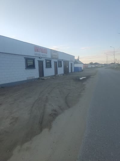 The image depicts a storefront in the city of Aral, Kazakhstan, under a clear, pale blue sky. The building is long and low, with a light-colored, ribbed metal roof and a facade made of light grey, rectangular blocks. Two signs are visible on the upper part of the facade. The left sign features large, pink, stylized Cyrillic lettering that reads "БУРЯТО" (Buryato), with smaller Cyrillic text below it reading "ШАРЫН МАРКЕТ" (Sharyn Market). To the right of this sign is another sign with more decorative script, also in Cyrillic, that appears to read "Гульфия" (Gulsfiya).
The storefront has several dark-colored doors and windows, some of which are barred with metal grates. The ground in front of the building is unpaved, consisting of dirt and sand with tire tracks and some puddles of water. To the right of the building, a paved road stretches into the distance, lined with scattered streetlights. In the background, several low-rise residential buildings and other structures can be seen, indicating a semi-urban or rural environment. The overall scene suggests an early morning or late evening setting due to the soft lighting. There are no people visible in the image.