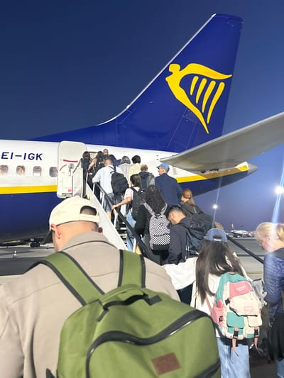 Passengers are boarding a Ryanair flight at night, likely in Luqa, Malta, given the aircraft registration EI-IGK. The passengers are ascending the boarding stairs towards the open door of the aircraft, with the tail fin visible showing the Ryanair logo. The focus is on the group entering the plane and the tail of the aircraft. People are seen wearing backpacks, and some are looking down as they climb the stairs. The environment suggests an airport tarmac under artificial lighting.
