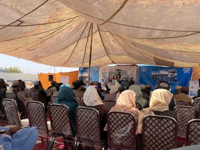 The image depicts an outdoor gathering under a large, light-colored fabric tent in Pir Dust, Afghanistan. Numerous individuals, predominantly appearing to be men and some women, are seated on patterned metal chairs arranged in rows on a red carpet. The attendees face forward towards a temporary stage or presentation area.
At the front, a speaker stands at a podium, addressing the audience. Behind the speaker, several vertical banners are visible, some displaying logos, text, and images related to infrastructure or construction, including depictions of bridges/roads and machinery. A blue banner with white imagery and text is prominent on the right side of the stage. A large, patterned orange and white fabric backdrop is also present. A black speaker on a stand is positioned on the left side of the stage.
The tent's fabric ceiling, supported by wooden poles and ropes, shows a faded floral or ornamental pattern. Beyond the tent's perimeter, a bright sky is visible, along with distant structures, fences, and sparse vegetation, indicating an open-air environment. The overall scene suggests a community meeting, official announcement, or public event.