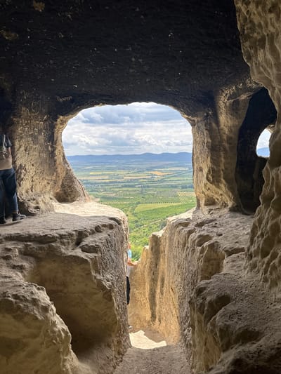 The image shows the view from inside a cave or rock shelter, looking out over a landscape. The cave entrance is roughly oval-shaped, framing a vista of green fields, rolling hills, and a sky with scattered clouds. The interior of the cave is rough, uneven rock, with variations in color from dark to light brown. A person is visible just outside the cave in the upper right of the frame, lying down on the rock. Lower in the frame, another person's legs can be seen. The location is near the village of Milanovo, Bulgaria.