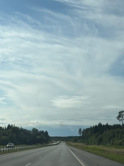 The image depicts a view of a straight highway in Rõmeda, Estonia, under a partly cloudy sky. The road, paved with asphalt, has two lanes in the visible direction of travel, marked with white dashed lines. A silver car is traveling in the left lane, and several other vehicles are visible further down the highway. On the left side of the road, a dense forest borders the highway, with a metal guardrail separating the road from the trees. On the right side of the road, there is a grassy verge, also bordered by trees. The sky is filled with wispy, white clouds against a light blue background. The overall scene is one of travel and nature, with the highway serving as a clear path through a forested landscape.