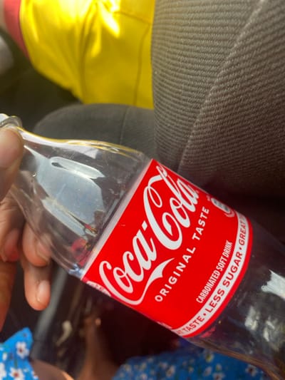 A human hand with dark skin tone holds a mostly empty, clear glass Coca-Cola bottle diagonally across the frame. The bottle features its standard red label, displaying "Coca-Cola ORIGINAL TASTE" and text indicating "CARBONATED SOFT DRINK" and "LESS SUGAR". The background consists of dark grey, ribbed fabric, characteristic of vehicle upholstery, occupying the upper right quadrant. A vibrant yellow fabric is partially visible in the upper left, while a blue fabric with a white floral pattern is visible in the lower left and bottom. The scene is situated in Unguwar Gaya, Nigeria.