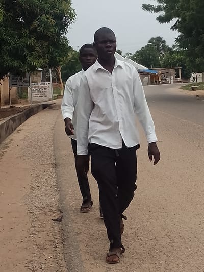 Two young men are walking on a paved road in Biliri, Nigeria. They are both wearing white, long-sleeved shirts and dark trousers, and sandals. The road is bordered by trees and a low wall. A sign for "Goade Sured Primary School Bonganje Biliri Lgea Gombe State Established 1927" is visible in the background. Other buildings and vegetation are seen further down the road.