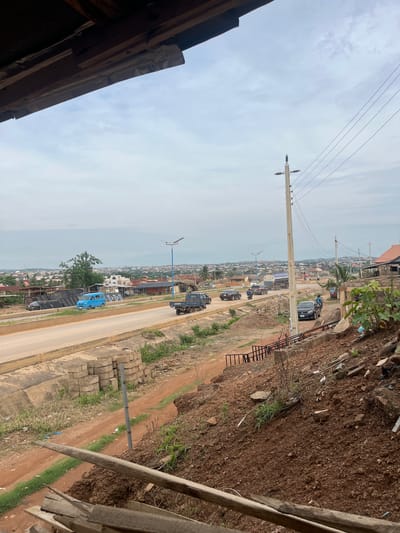 An elevated, wide-angle image captures a semi-urban landscape in Offatedo, Nigeria, under a predominantly overcast sky. The photograph is taken from a vantage point looking down onto a main road that stretches horizontally across the mid-ground.
In the immediate foreground, on the right side, is a steep, reddish-brown dirt embankment, appearing rugged with sparse greenery, scattered debris, and partial wooden structures. On the far left, the edge of a building's dark roof frames the shot, indicating the camera's elevated position.
A prominent concrete utility pole stands near the left side of the road, supporting multiple power lines that crisscross the cloudy sky. Further down the road, another smaller, blue-colored pole with a horizontal arm, possibly for street lighting or signage, is visible.
The paved road features a yellow median line and accommodates several vehicles. A dark blue truck with the partial text "TOYOTA" on its side is in the right lane, followed by a dark-colored sedan. In the opposite, left lane, a light blue minibus is present, and further ahead, other vehicles and a motorcycle can be seen. Along the dirt verges flanking the road, stacks of grey concrete blocks are neatly arranged.
The background reveals a sprawling settlement with numerous buildings, most appearing to be single-story houses with varied roof colors, extending towards the hazy horizon. The scene depicts a typical daytime environment, suggesting a mix of residential activity and ongoing development in the area.