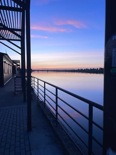 The image depicts a twilight scene by a river in Sovetsk, Russia, likely near the railway bridge that spans the Neman River. In the foreground, a dark metal railing runs along a walkway paved with dark bricks and concrete. To the left, a dark building with closed shutters and a slatted roof structure is visible. The river itself is calm and reflects the soft colors of the sky, which transitions from a deep blue at the top to hues of pink, orange, and light yellow near the horizon. A silhouette of a long railway bridge with intricate metalwork is visible across the river, and beyond it, a treeline marks the opposite bank. The overall atmosphere is peaceful and serene, characteristic of a sunset or sunrise.
