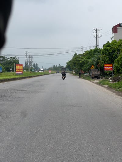 The image depicts a scene on a paved road in Nam Viêm, Vietnam, under an overcast sky. The road has visible patches of wear and tear. On the left side of the road, there is a grassy verge with a red sign that reads "NHÀ NGHỈ HUỲNH HƯƠNG" with an arrow pointing to the right, and further down the road, a white sign with a red border and a red triangle. Power lines and utility poles are present in the background, along with a cluster of trees. On the right side of the road, there is a white building with a red roof, a utility pole, and more trees. Vehicles are visible on the road, including a truck, a motorcycle with a rider, and a person on a bicycle. Another red sign with similar text to the one on the left is visible on the right. There is a parked truck on the right side of the road.