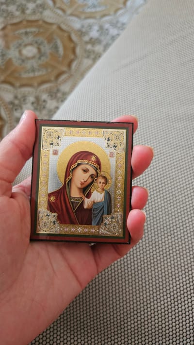 A hand holds a small, rectangular icon depicting the Virgin Mary and the infant Jesus. The icon is framed by gold ornamentation and features Greek lettering. The background consists of a textured surface, possibly a sofa, and a blurred patterned floor. The image was captured in Mondolfo, Italy.