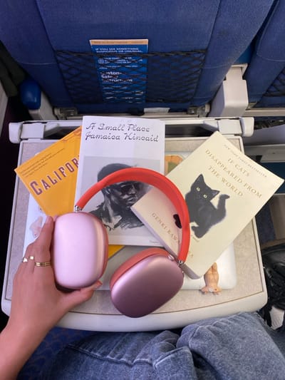 The scene depicts a passenger's perspective inside an airplane cabin. The specific location in Oceanside, United States, cannot be determined from the image, as the environment is an airplane interior.
In the foreground, a light-colored, deployed airplane tray table is visible. Resting on the tray table are three books and a pair of headphones. The books include an orange-covered book partially showing "CALIFORNIA," a white-covered book titled "A Small Place" by Jamaica Kincaid featuring a black and white image of a man in sunglasses, and a cream-colored book titled "If Cats Disappeared from the World" by Genki Kawamura, displaying a black cat on its cover. A small, light-colored figure is partially visible beneath the "If Cats Disappeared from the World" book.
A person's left hand, wearing two gold-colored rings on the ring finger, is holding a pair of rose gold/pink Apple AirPods Max headphones with an orange/coral fabric headband. The individual's lap, covered by blue denim jeans, is visible in the lower right portion of the frame.
In the background, a blue fabric airplane seat with a black mesh seatback pocket is visible. The pocket contains various white and blue informational pamphlets, including safety instructions and a card with partially legible text such as "WE'RE ALL IN THIS TOGETHER" and "IF YOU SEE SOMETHING SUSPICIOUS OR UNUSUAL." The overall environment is consistent with a commercial airline passenger seat.