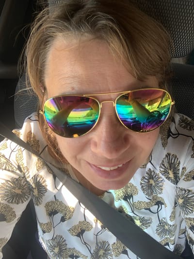 The image shows a close-up selfie of a woman wearing sunglasses with rainbow lenses and a white shirt with a floral pattern. She is inside a vehicle, indicated by the seatbelt and the reflection in her sunglasses, which shows the dashboard and a road scene. The woman is smiling and looking towards the camera. The setting appears to be daytime, with sunlight illuminating her face. No specific landmarks or details definitively place the image in Азерскі сельскі Савет, Belarus.