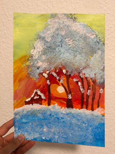 This is a close-up, horizontal shot of a painting held in a person's hand. The painting depicts a winter scene with a sunset. The left side of the painting features a large, abstract white and light blue mass, possibly representing snow or a snow-covered tree. The right side of the painting is dominated by a vibrant sunset with streaks of orange, yellow, and red, suggesting a warm glow. Dark brown branches with white accents, resembling blossoms or snow, extend across the canvas, overlapping both the snowy and sunset areas. A small, circular white shape in the center of the sunset area likely represents the sun. A small, dark brown structure with white accents, possibly a house, is visible on the lower right side. The background on the far left is a light green, while the far right is a solid blue. The painting is rendered in an impressionistic style with visible brushstrokes. The overall mood is one of contrast between cold and warmth, winter and sunset. There is no visible text in the image.