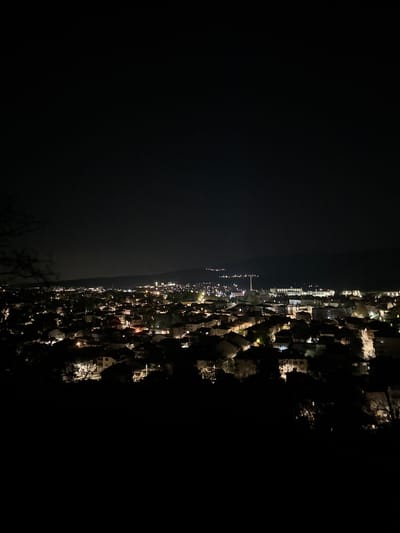 A nighttime view from Sveti Konstantin, Bulgaria shows an area of illuminated buildings and streetlights along the coastline. To the left of the image is a dark body of water, possibly the Black Sea. The lit-up area suggests a residential or commercial district with various buildings and structures, some with visible architectural details. The dark areas surrounding the lit-up zones likely represent undeveloped land, natural terrain, or shadows cast by buildings. The overall scene captures the urbanized and naturally dark environment of Sveti Konstantin at night.