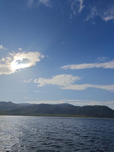 The image depicts a serene natural landscape, likely a body of water bordered by rolling hills under a bright blue sky with scattered clouds. The sun is visible, peeking through a formation of clouds, creating a subtle halo effect. The water in the foreground shows gentle ripples, reflecting the light. The hills in the background are covered in vegetation, with a treeline along their crests. The overall impression is one of peacefulness and natural beauty. The location is identified as the city of Manat, Kazakhstan.