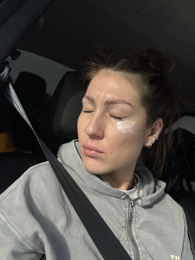 A female subject is depicted from the mid-chest upward, seated in what appears to be the front passenger seat of a vehicle, secured by a dark seatbelt across her torso. Her eyes are closed, and her head is tilted slightly to her right, with direct sunlight illuminating the left side of her face. A dollop of white cream or lotion is visible on her left cheekbone, below the outer corner of her left eye. She has dark hair pulled into a messy bun and wears a light grey zippered hoodie over a white undershirt with a thin silver chain visible at her neck. Her left ear features multiple piercings, including various studs and rings, one shaped like a flower. The vehicle's interior appears dark, with a black headrest directly behind her. No specific location within Ustinovo, Russia, or any other city, can be determined from the visual evidence presented in the image.