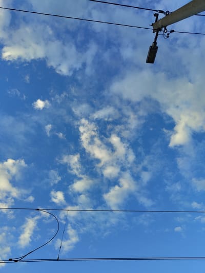 The image shows a bright blue sky with scattered white clouds. In the upper left quadrant, a streetlight is mounted on a pole, connected by wires. These wires, along with others, run vertically through the frame, suggesting an outdoor, possibly urban or suburban, setting. The lighting indicates it is daytime, with the sun illuminating the clouds. No people are visible, and no specific activity is taking place. The image captures a serene moment of a cloudy sky with man-made infrastructure. The location context provided is Цибино, Russia.