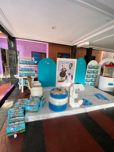 The image shows a birthday party setup in a building located at 4.048 latitude, 9.218 longitude in Bonadikombo, Cameroon. There are numerous party favors, all with a blue and white color scheme, and a blue and white theme that is consistent with the birthday decorations. The party favors have the name "Joel-Ethan" written on them. The scene includes several objects, including a white table with a white cloth on top, a white chair with a gold trim, a blue and white stool, and a white wall with a poster that says "Happy Birthday Joel-Ethan". The poster features a young boy sitting on a red toy car. The room has a white ceiling, and the walls are painted pink and brown. The floor is red and black. The building appears to be part of a larger complex. The image was likely captured during a birthday party for a child named Joel-Ethan.