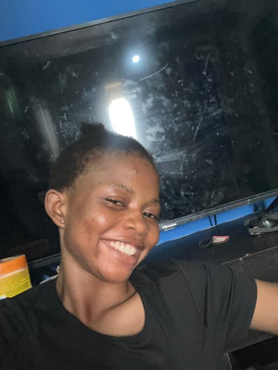 A person with short, dark hair and an earring is smiling at the camera. The person is wearing a black top. The background includes a large, dark, reflective surface, possibly a television screen, and a portion of a blue wall. A yellow package is visible near the bottom right corner. The location is unknown.