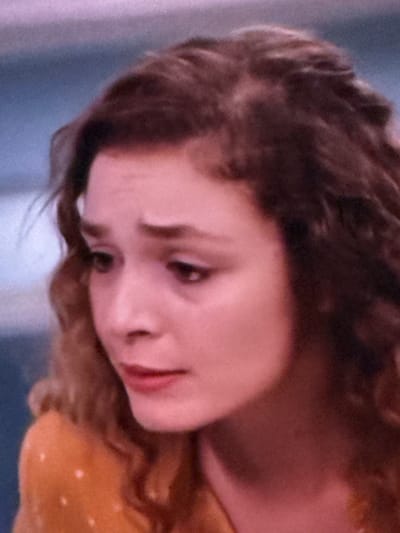The image is a close-up, medium shot of a young woman's head and shoulders, angled from the left. She has wavy, dark brown hair that frames her face. Her skin tone is fair, with a slight pinkish hue. Her eyes are downcast and appear to be a dark brown. Her eyebrows are arched, and she has a slight frown. Her nose is small and slightly upturned. Her lips are full and painted with a muted red lipstick. She is wearing an orange top with a subtle polka dot pattern. The background is blurred and appears to be a light blueish-gray. There is no discernible location in the city of Springboro, United States of America visible in the image.