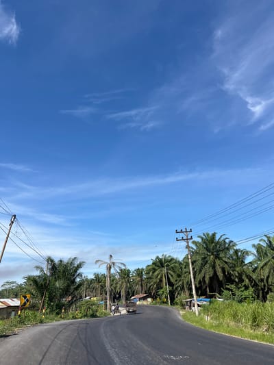 The image shows a roadside scene in Bukitrata, Indonesia, under a clear blue sky with wispy white clouds. The perspective is from a low angle looking upwards and to the right. To the right of the frame, a paved road curves away, bordered by lush green vegetation and numerous palm trees. A power line runs diagonally across the upper portion of the image, supported by a wooden utility pole.

Along the roadside, there are several small structures and some people. In the distance, a few vehicles, including a small truck, are visible. A yellow diamond-shaped sign with a black cross is placed by the roadside. The overall impression is a rural or semi-rural setting on a bright, sunny day. The time of day appears to be late morning or early afternoon, given the strong sunlight and shadows. There is no discernible text in the image.