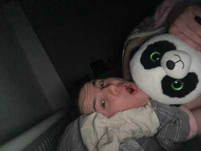 The image depicts a dimly lit interior scene, likely a bedroom, located in Benningen, Germany. An individual with light skin is shown lying diagonally across the frame, with their head positioned towards the lower left. Their head rests on a light-colored pillow, partially covered by a grey and white striped blanket. The individual's mouth is open in an 'O' shape, and their eyes are open, directed towards the camera. A large, white and black panda plush toy with prominent green glittery eyes is positioned adjacent to the individual's head, appearing to be held by their right arm, which is partially visible. The visible attire suggests light-colored or patterned sleepwear. The background is predominantly dark, with a vertical white object, consistent with a radiator, visible along the far-left edge of the frame.
