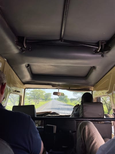 The image captures the interior of a vehicle, looking forward through the windshield, offering a view of a road and surrounding landscape. The scene is set in Kwa Kuchinia, Tanzania.

Inside the vehicle, the walls are upholstered with dark, possibly padded material. On the left side, a sturdy black metal bar runs horizontally, with a vertical support connected to it, suggesting a robust interior design, possibly for passenger stability or gear. The overall impression is that of a utility or safari-style vehicle.

Through the front windshield, a paved road with a single white dividing line is visible, stretching straight into the distance. Lush green trees line both sides of the road, indicating a natural or rural environment. The sky appears clear and bright, suggesting daytime with fair weather conditions. A rearview mirror is visible in the upper center of the windshield area.

Two individuals are partially visible within the vehicle. In the front, likely the driver, a person's head and upper body are seen, wearing a dark top. Another person's lower body, including their legs dressed in light-colored pants, is visible to the right, suggesting they are seated in the front passenger area. Further to the lower right, another person's legs and feet, possibly belonging to a passenger in the back, are visible.

The activity depicted is travel by vehicle along a road. No specific event or strong emotions are discernible. There is no visible text in the image. The bright natural light indicates it is daytime.