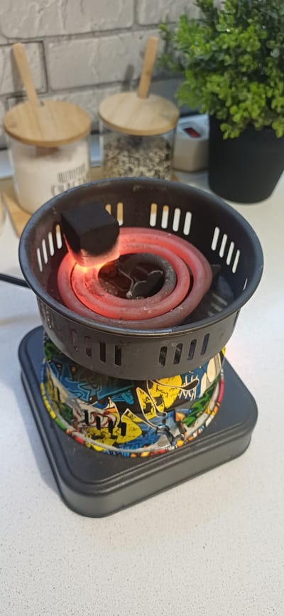 The image depicts a close-up view of an electric hookah charcoal burner. A single black charcoal cube, glowing red hot on one side, rests on the coiled heating element. The burner itself is black with slotted sides. It sits atop a colorful, graffiti-themed decorative sleeve. Below the sleeve is the black base of the burner, which has a square footprint. The background is blurred but suggests a kitchen or countertop setting with two glass jars with wooden lids, one containing a white substance and the other a granular material. A green artificial plant in a black pot is also visible to the right. The scene is well-lit. There is no indication of Zuevka, Russia, in the image.