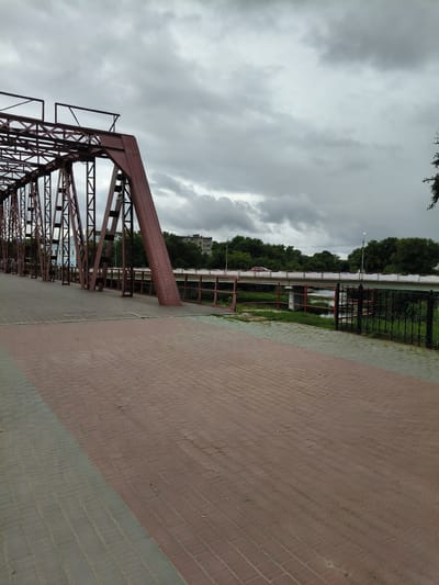 The image shows a pedestrian walkway on a bridge made of red steel trusses. The walkway is paved with reddish-brown and gray rectangular bricks. To the right of the walkway, there is a black ornamental metal fence and lush green grass. Beyond the fence, a river flows under a modern concrete bridge, on which a red car is visible. The riverbanks are lined with dense green trees. In the background, several buildings and more trees are visible under a cloudy sky. The bridge structure appears to be a historic metal truss bridge, possibly a pedestrian or vehicle bridge that has been repurposed. The overall scene suggests an urban environment with a mix of old and new infrastructure, likely located near a body of water.
