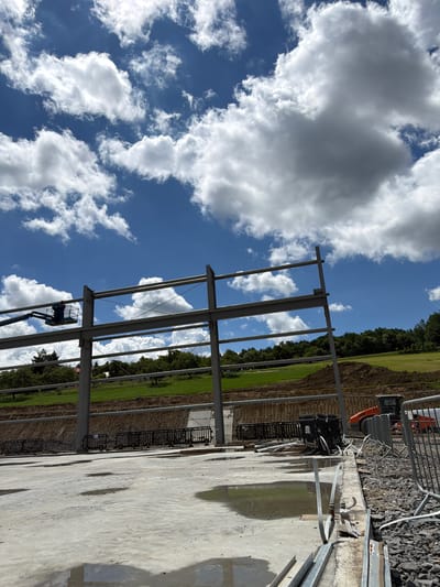 The image depicts a construction site in Skrzydlna, Poland, under a partly cloudy sky. A large steel frame structure, likely for a building, dominates the midground. In the left midground, a worker is visible in a cherry picker, operating on the steel frame. In the background, a grassy hillside with trees borders the site, and a pile of excavated earth is present. In the right midground, an orange construction vehicle, possibly a roller, is parked near a temporary fence. The foreground consists of a wet concrete surface with puddles, scattered construction materials, and more temporary fencing.