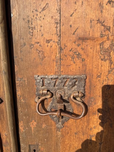 The image shows a close-up of an old wooden door, likely in Tossa de Mar, Spain, given the style of the door hardware. The door is made of weathered wooden planks, showing signs of age with cracks, peeling paint, and a rough texture. Mounted centrally on the door is a heavy, ornate metal door knocker and escutcheon plate. The metalwork is dark and appears to be cast iron, featuring decorative scrollwork and what appears to be the year "1773" inscribed in raised numerals on the escutcheon. The knocker itself is a ring attached to a decorative handle. A keyhole is visible below the knocker. The sunlight casts a strong shadow to the right of the door knocker, obscuring part of the wood's texture and surface detail. A vertical wooden element, possibly part of the door frame or another architectural feature, is visible on the left side of the image.
