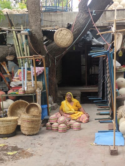 In Jodhpur, India, a woman in a bright yellow sari and patterned skirt sits cross-legged on a concrete ground. She is surrounded by an assortment of woven bamboo baskets. To her right, several small, lidded baskets with pink accents are neatly arranged. Behind her, a large stack of taller baskets forms a prominent display. In front of the stack of baskets, a very large, wide basket rests on what appears to be a spare tire. To the left of the large stack, a round, flat basket is also visible. The setting appears to be an outdoor market or workshop. A large tree with rough bark dominates the background, with its branches extending upwards and secured with ropes. Various bamboo poles, some bundled together, are propped against the tree and a wooden structure. A blue plastic bin is visible to the left of the basket display. The ground is stained with water and scattered with dry yellow leaves. In the background, there is a building with dark openings and metal shelving units filled with more items. Several large, round, pale objects are stacked against the wall in the right background, some covered by a blue tarp. The overall scene depicts a vendor in an outdoor environment surrounded by her merchandise.