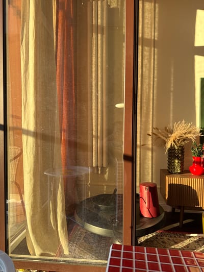 The image is an outdoor shot of a store window. The window is reflecting the interior of the store, as well as the outdoors. Inside the store, there are shelves with various decorative items, including a red vase, a green glass vase filled with dried flowers, and other smaller decorative objects. There is also a red cylindrical object visible on a shelf. The store interior appears to be well-lit, with natural light casting shadows. Outside the window, there are blinds or curtains visible, in shades of beige and brown, suggesting a cozy interior. The lighting suggests it is daytime, with sunlight creating strong shadows, indicating a sunny day. The reflection in the window shows glimpses of an outdoor environment, possibly a street or public space. The overall scene is one of a well-decorated retail space. There is no visible text.