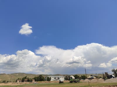 The image captures a construction site in Boroldoy, Kyrgyzstan. A bright blue sky with scattered white clouds dominates the upper portion of the frame. In the middle ground, a white building with large windows is visible, flanked by trees. A green cement mixer is positioned to the right of the building, with its chute extended, suggesting ongoing work. Piles of gravel and sand are present, along with a smaller, white tank-like vehicle. In the background, rolling hills covered in dry grass stretch to the horizon. The foreground consists of a grassy field. The scene appears to be devoid of people.