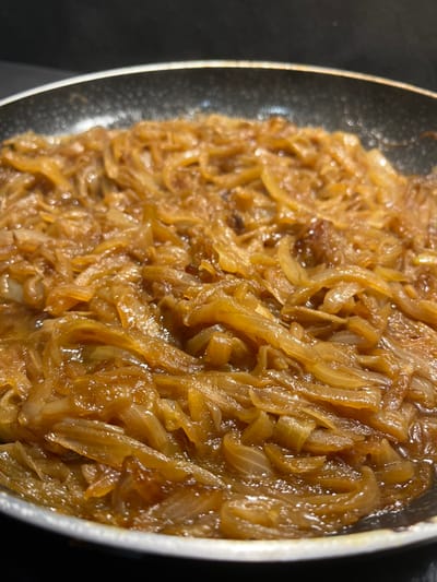 A close-up image displays a frying pan filled with thoroughly caramelized, golden-brown sliced onions. The onions are uniformly cut and exhibit a glossy sheen from moisture or cooking fat. The pan features a dark, speckled non-stick interior and a metallic rim. The background is dark and out of focus, consistent with an indoor kitchen setting. This scene is located in Foetz, Luxembourg. No individuals, actions, or interactions are visible.