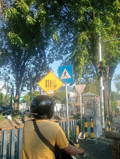 The image depicts a street scene in Banjarkemantren, Indonesia, with a focus on a person riding a motorcycle. The rider, seen from behind, wears a black helmet and a yellow t-shirt. The motorcycle has a "HONDA" logo visible on the helmet. In the foreground, a black railing separates the roadway from what appears to be a railway track.

Several traffic signs are present. A yellow diamond-shaped sign with a symbol of a railroad crossing gate is prominent. Adjacent to it is a blue square sign with a white pedestrian symbol, indicating a pedestrian crossing. A triangular yield sign is also visible.

A traffic light pole with red and black lights stands near the signs. Behind the signs and traffic light, trees with lush green foliage provide shade. In the background, buildings and other vehicles, including a black car and a motorcycle with a rider, suggest an urban environment. The ground is a mix of asphalt and dirt, with some debris. The overall lighting indicates it is daytime, likely morning or afternoon, with sunlight filtering through the trees.
