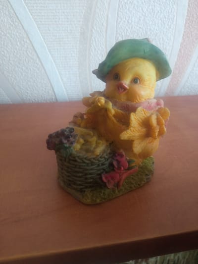 A ceramic figurine of a yellow chick wearing a green hat and a pink scarf sits in a woven basket on a brown wooden surface. The background features a textured white wall with vertical decorative elements. The figurine is decorated with small pink flowers.