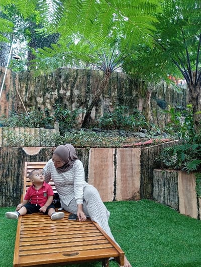 The image depicts an outdoor garden area, located in Kuwukan, Indonesia. An adult female and a young child are seated on a light-colored wooden slatted lounge chair positioned on green artificial turf. The adult female, wearing a light brown hijab, a white and light grey horizontally striped long-sleeve top, and light grey textured pants, sits with her left leg extended along the chair. Her gaze is directed towards the child. A dark-colored smartphone rests on the chair beside her right hip. The child, an infant or toddler male, wearing a red short-sleeve polo shirt, black shorts, and white shoes, sits to the left of the female, looking upwards at her. Behind them, a tiered, weathered concrete wall with sections resembling tree trunks forms the background. Various green plants and multiple tree ferns with large fronds are visible, both along the wall's base and extending into the upper portion of the frame.