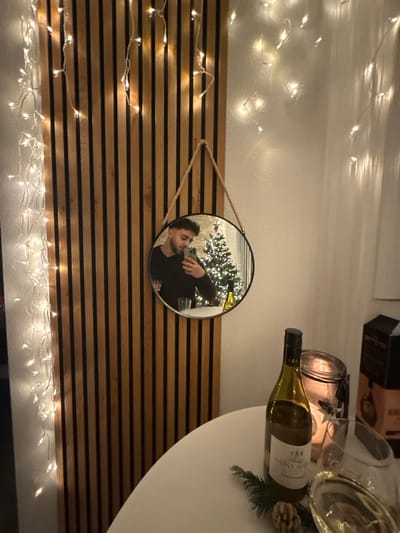 A horizontal, wood-slatted wall is adorned with a circular mirror. The reflection in the mirror shows a man in a black shirt holding a phone, capturing a Christmas tree in the background. Strands of twinkling fairy lights frame the top and bottom of the wall. To the right, a white table displays a bottle of white wine, a candle, a glass, and pine boughs. Above these items, blurred boxes are visible against a white wall.