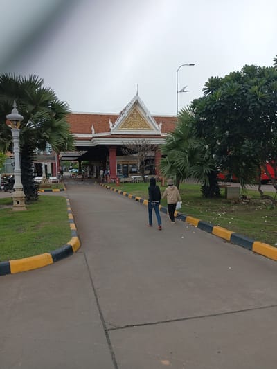 The image depicts the Bavet border crossing in Bavet Quarter, Cambodia, under an overcast sky. The central focus is a large building with a traditional tiled roof featuring ornate gold detailing, likely serving as a customs or immigration checkpoint. Two individuals, one wearing a black hooded jacket and jeans, and another in a tan jacket and dark pants, are walking away from the camera on a paved road leading towards the building. The road is bordered by green grass and decorative curbs painted yellow and black. To the left, a tall lamppost stands near a palm tree, and to the right, a mature tree with green foliage and white flowers is visible, along with a red bus parked in the background. Several other people are seen near the building, suggesting ongoing activity at the crossing. The scene is captured from a medium distance, showing the approach to the border facilities.