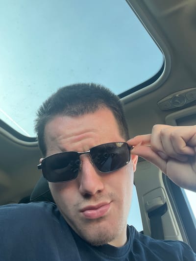 A selfie was taken inside a car, presumably in Benningen, Germany. The man is holding sunglasses in front of his face. The car has a sunroof, through which clear skies are visible. The interior ceiling is beige. The man has a medium complexion, with stubble and short dark hair. He is wearing a dark-colored shirt. The car's interior includes controls on the ceiling.