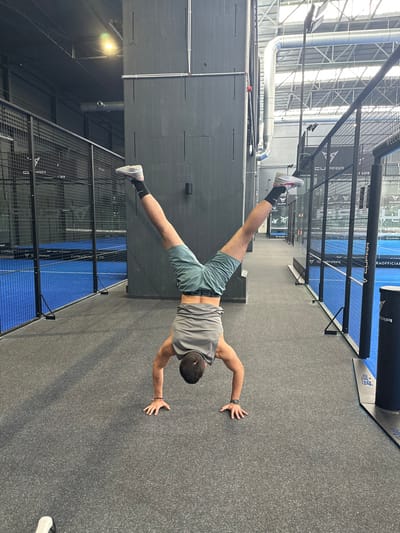 An individual is depicted performing a handstand on a dark grey, textured floor within the Padel Base Mülheim facility, located at Kiebitzweg 30-32, 45479 Mülheim an der Ruhr, Germany. The male subject, viewed from behind, wears a grey sleeveless tank top, dark green shorts, black ankle socks, and light-colored athletic shoes. His legs are spread overhead in a 'V' shape, and his hands are placed firmly on the floor. Tattoos are visible on both forearms, and a dark watch is on his left wrist. The immediate surroundings feature a prominent dark grey industrial pillar or wall to the subject's left and a padel court to his right. Two padel courts are visible in the background, characterized by bright blue playing surfaces enclosed by black metal mesh fencing and transparent glass panels, displaying "CUPRA OFFICIAL" and "CUPRA PADELCLUB.DE" branding. An upright dark cylinder with "CUPRA" branding stands near the right court. The facility features a high industrial ceiling with exposed silver ductwork, structural beams, and artificial lighting.