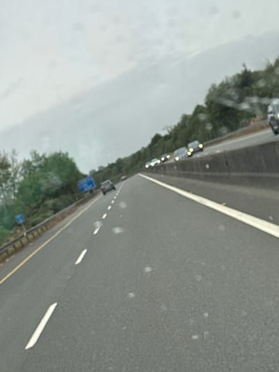 The image depicts a view from a vehicle traveling on a highway, likely the M11, in or near Gorey, Ireland. The highway has multiple lanes marked by white lines, with a concrete barrier separating lanes from the opposing direction of traffic. On the left side of the road, a metal guardrail is visible, with trees and foliage beyond it. Blue highway signs are also present on the left. A line of cars is seen in the distance, traveling in the same direction as the viewer's vehicle. The sky is overcast, suggesting a cloudy or possibly rainy day. The road surface appears wet, with small white specks possibly indicating water droplets or reflections on the windshield.