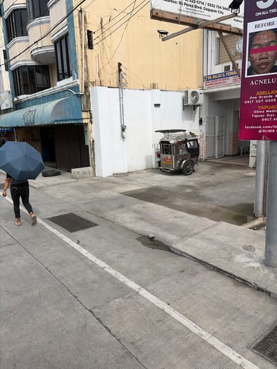 A person carrying a blue umbrella walks on a sidewalk in Dagupan City, Philippines. They are wearing black pants and beige sandals. Behind them, a three-wheeled vehicle, known as a tricycle, is parked. The scene includes a building with a yellow facade, a blue awning, and several windows. A sign for "DR. MALOU S. FERNAN" is visible on the building, along with advertisements for medical services. Power lines and utility poles are present in the background. The ground is paved with concrete, showing some signs of wear and tear, including cracks and water stains. The lighting suggests it is daytime.