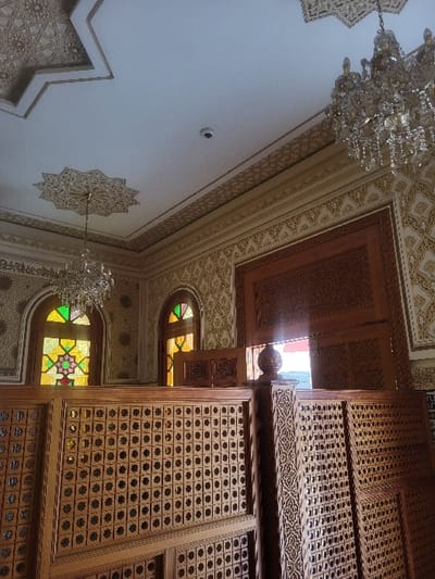An interior scene in Oulad Hcine, Morocco, depicts a decorated room featuring traditional Moroccan architectural elements. The white ceiling displays elaborate plasterwork, including a large multi-pointed star motif and linear geometric patterns. Two large, multi-tiered crystal chandeliers are suspended from the ceiling. A small, circular security camera is mounted centrally on the white ceiling. The walls are covered in repeating gold and tan geometric and floral patterns. Two arched windows on the left feature colorful stained-glass panels with geometric designs. Extensive light-colored wooden elements are present, forming partitions and panels carved with intricate geometric patterns, specifically small squares containing star shapes. Some of these wooden panels appear to be stacked or leaning on the right. A large, dark wooden archway or doorframe with carved details is visible on the right wall, emitting bright light from beyond. No individuals are present.
