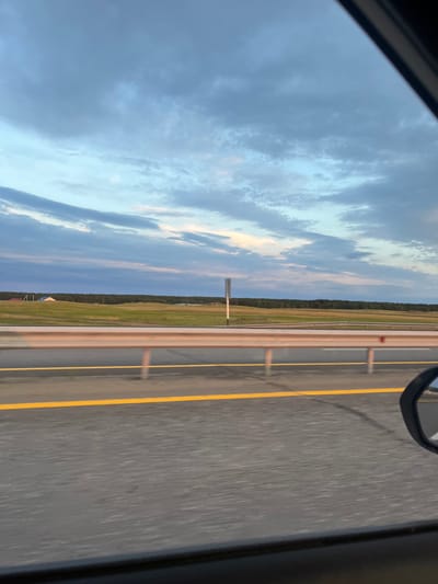 The image depicts a view from a moving vehicle, likely on a highway, looking out towards a rural landscape under a cloudy sky. The foreground shows a section of asphalt road with a bright yellow line marking the lane. A metal guardrail separates the road from the grassy fields beyond. In the middle ground, rolling green hills are dotted with what appear to be a few buildings, including one with a distinctive white, pyramid-shaped roof. A dark line of trees forms a horizon across the mid-distance. The sky is filled with a mixture of light blue and grey clouds, suggesting either early morning or late evening. The reflection of the sky and clouds is visible in the side-view mirror on the right side of the frame. The overall impression is one of travel through a serene, expansive countryside. The specific location in Алтынды ауылдық округі, Kazakhstan is not visually identifiable within the image itself.
