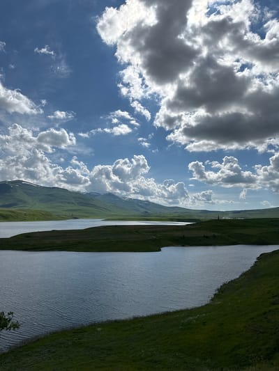 The image depicts a serene landscape with a large body of water in the foreground, reflecting the dramatic, cloud-filled sky. Rolling green hills, some with patches of snow, stretch across the horizon. The sky is a vibrant blue, punctuated by numerous fluffy white clouds and darker, more dramatic storm clouds, creating a play of light and shadow across the scene. A dark green, grassy slope dominates the right foreground, meeting the edge of the water. The scene is devoid of any people or structures, suggesting a remote, natural environment. Based on the description, this landscape could be the vicinity of Tsghuk, Armenia, known for its natural beauty and mountainous terrain.
