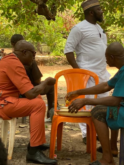 The image displays an outdoor scene in Ochadam, Nigeria, under a canopy of green trees with a visible branch in the upper left. The ground is composed of dry, reddish-brown earth with scattered leaves. Four adult males are present in the foreground and midground. On the left, a bald man wearing dark sunglasses, an orange traditional tunic, and matching trousers sits on a white plastic chair, facing right. His left arm is bent, and his hand is clenched; he wears black dress shoes. Behind him, another man is partially visible, wearing a dark top. In the center, a bearded man in a white traditional tunic and a striped brown and black cap stands, facing right, with his left hand in his pocket. To the right, another bald man, wearing a teal traditional top and patterned dark shorts, is seated. He is handling yellowish food from a white, oval-shaped tray placed on an orange plastic chair, which functions as a table. Sunlight filters through the overhead foliage.