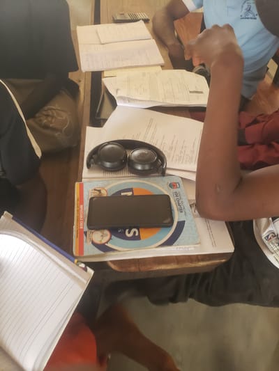 The scene depicts a desk cluttered with academic materials. There are several pages of notes and calculations, a notebook, and a textbook with the title "Federal Polytechnic Ede Chapter," indicating the location is likely Ede, Osun State, Nigeria. A black smartphone rests on the textbook. A pair of JBL headphones also sits on the desk. Two people are partially visible: one wearing a light blue shirt and another wearing gray pants and a shirt with an NBA logo. The subjects appear to be studying or working at the desk.
